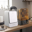 Phrozen Air Purifier Max connected via exhaust hose beside a Sonic Mighty Revo 16K resin printer on a workbench, with printed miniatures, resin bottles and cleaning supplies nearby.