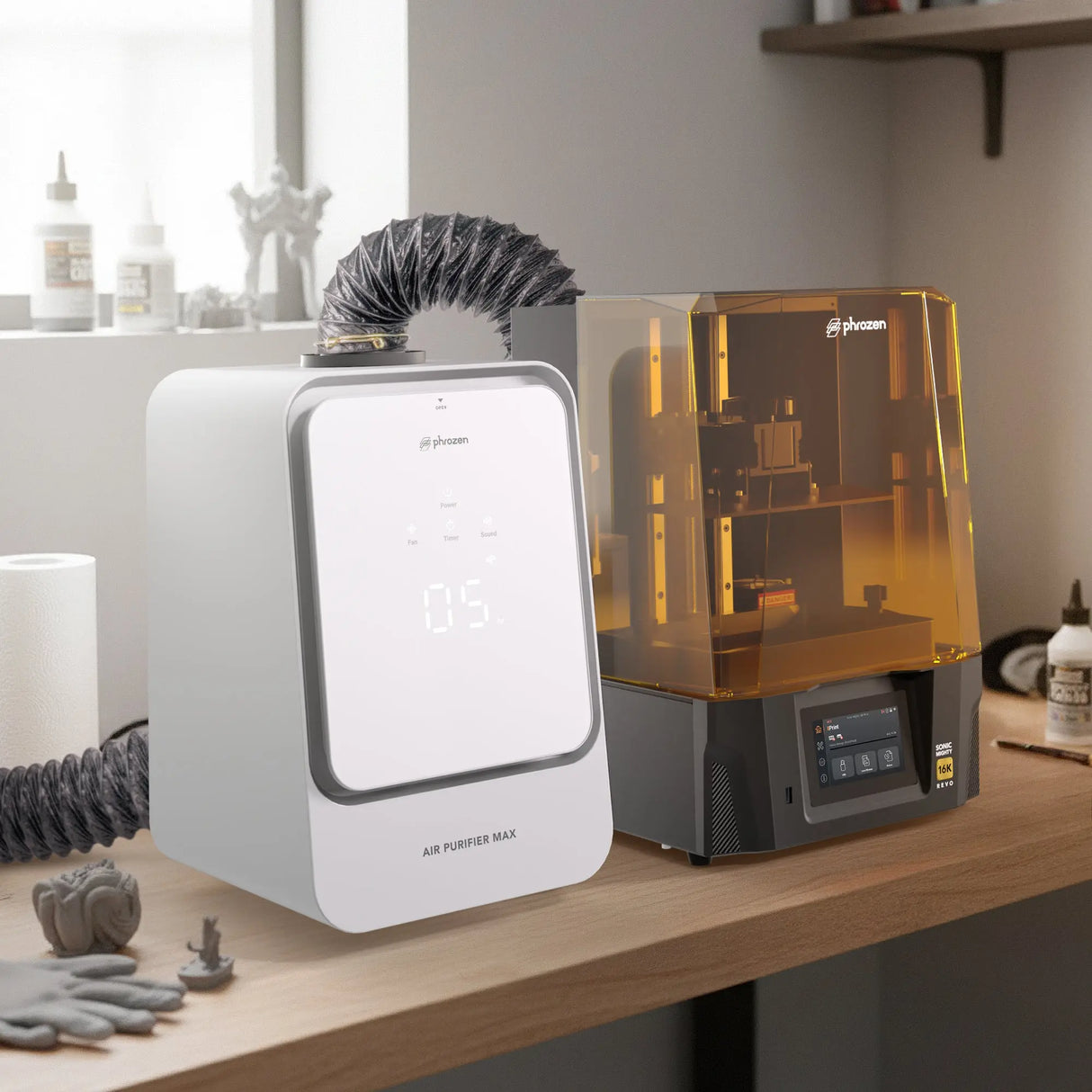 Phrozen Air Purifier Max connected via exhaust hose beside a Sonic Mighty Revo 16K resin printer on a workbench, with printed miniatures, resin bottles and cleaning supplies nearby.